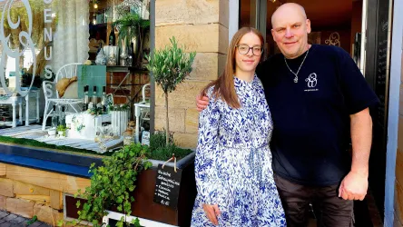 Ein Mann und eine Frau stehen lächelnd vor einem Ladengeschäft. Im Hintergrund sind Pflanzen und Möbel sichtbar, die zur Dekoration des Geschäfts beitragen.