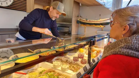 Ein Verkäufer reicht einer älteren Dame eine Auswahl an frischen Speisen in einem Marktstand. Der Stand ist hell und einladend, mit vielen verschiedenen Lebensmitteloptionen.