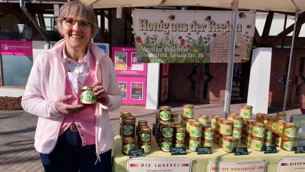 Eine Frau steht an einem Stand und hält ein Glas Honig in der Hand. Im Hintergrund sind viele Honiggläser aufgereiht, und ein Schild zeigt "Honig aus der Region".