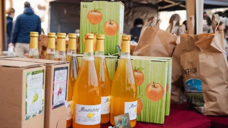 Eine Auswahl an Flaschen mit Apfelsaft und einigen Pappkartons auf einem Markt. Die Flaschen sind leuchtend orange und die Kartons zeigen frische Äpfel.