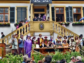 Eine Gruppe von Menschen in historischen Kostümen steht auf einer Treppe vor einem Gebäude. Einige Personen halten Holzstäbe und es gibt eine festliche Atmosphäre.