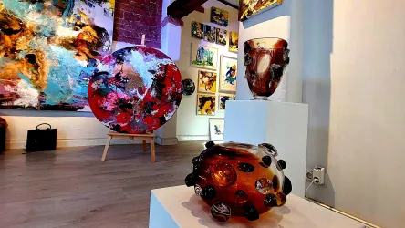 Eine moderne Kunstgalerie mit bunten Glaskunstwerken auf einem weißen Tisch. Im Hintergrund sind große, farbenfrohe Gemälde und Skulpturen sichtbar.