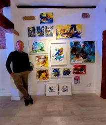 Ein Mann steht neben einer Wand mit mehreren farbenfrohen Gemälden. Die Kunstwerke zeigen verschiedene abstrakte Designs und sind in unterschiedlichen Rahmen präsentiert.