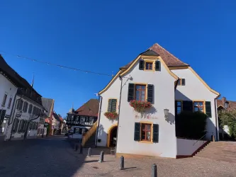 Ein charmantes, weißes Gebäude mit bunten Fensterläden und Blumenkästen. Die Straße ist ruhig und von traditioneller Architektur umgeben, und der Himmel ist strahlend blau.