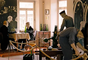 Eine historische Ausstellung mit zwei Figuren an einem Tisch und einer dritten Figur, die eine Flasche abfüllt. Die Wände sind mit Bildern von früheren Zeiten dekoriert.