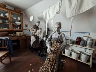 Ein historisches Kücheninnere mit zwei Figuren, die traditionelle Aufgaben erledigen. Im Hintergrund sind Holzmöbel und verschiedene Küchenutensilien zu sehen.