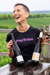 Eine fröhliche Frau hält zwei Weinflaschen in den Händen und lächelt. Im Hintergrund sind grüne Weinberge zu sehen.