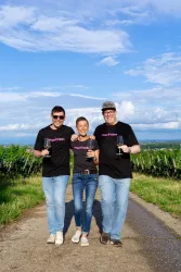Drei Personen stehen lächelnd auf einem Schotterweg zwischen Weinreben. Sie halten Gläser in der Hand und tragen schwarze T-Shirts mit einem Logo.