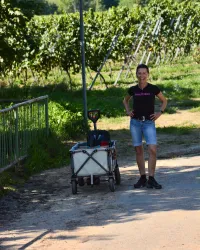 Eine Frau steht neben einem kleinen Wagen auf einem Weg zwischen Weinreben. Sie trägt ein schwarzes T-Shirt und Shorts und lächelt in die Kamera.