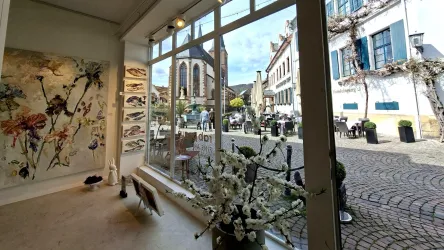 Ein Kunstgeschäft mit großen Fenstern, die auf einen malerischen Platz blicken. Im Hintergrund sind eine Kirche und Cafés sichtbar, umgeben von charmanten Gebäuden.