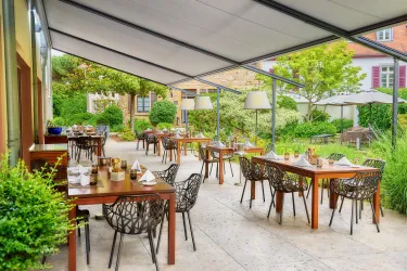 Ein gemütlicher Außenbereich eines Restaurants mit Holzstühlen und Tischen. Umgeben von grünen Pflanzen und Sonnenschirmen.