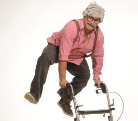 Ein älterer Mann mit einer Perücke springt über einen Rollator. Er trägt ein rotkariertes Hemd und hat einen lustigen Gesichtsausdruck.