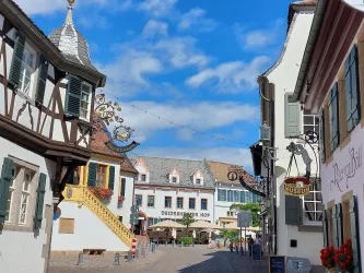 Eine malerische Gasse mit traditionellen Fachwerkhäusern und einem einladenden Restaurant im Hintergrund. Der Himmel ist blau mit wenigen Wolken, was eine angenehme Atmosphäre schafft.