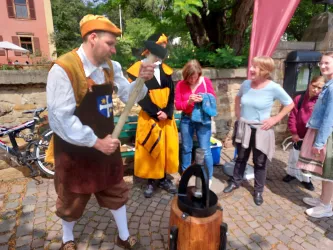 Ein Mann in historischer Kleidung zeigt eine traditionelle Handwerkskunst. Eine Gruppe von Zuschauern beobachtet interessiert.
