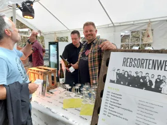 Eine Weinverkostung in einem Zelt mit zwei Männern, die Wein einschenken. Im Hintergrund sind Gläser und eine Tafel mit Informationen über die Weinsorten zu sehen.