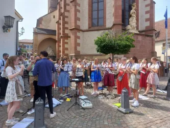 Eine Gruppe von Musikanten spielt an einem Platz vor einer Kirche. Sie tragen bunte Kleider und genießen das sonnige Wetter.