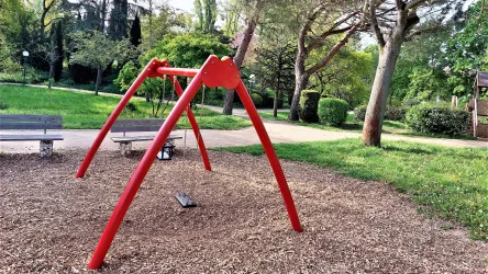 Ein Spielplatz mit einer roten Schaukel steht auf einem mit Holzspänen bedeckten Boden. Im Hintergrund sind Bänke und grüne Bäume zu sehen.