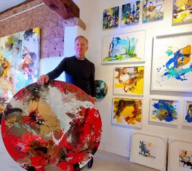 Ein Künstler steht in einer Galerie und hält ein rundes, buntes Gemälde. Im Hintergrund sind mehrere weitere farbenfrohe Kunstwerke an der Wand zu sehen.