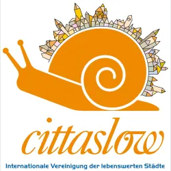 Das Bild zeigt ein stilisiertes, orangefarbenes Schneckenlogo mit einer bunten Häuserlandschaft auf dem Rücken. Darunter steht der Schriftzug "cittaslow" und der Zusatz "Internationale Vereinigung der lebenswerten Städte".