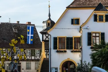 Ein historisches Gebäude mit gelben Fensterrahmen und einem hübschen Dach ist zu sehen. Im Hintergrund hängt eine Flagge mit blauem und gelbem Design.