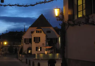 Ein charmantes Gebäude in der Dämmerung mit warmem Licht von einer Laterne. Im Vordergrund ist ein Brunnen zu sehen, umgeben von einer ruhigen Atmosphäre.