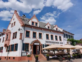 Ein historisches Gebäude mit dem Namen "Deidesheimer Hof". Die Fassade ist hell, und Tische stehen unter Sonnenschirmen im Freien.