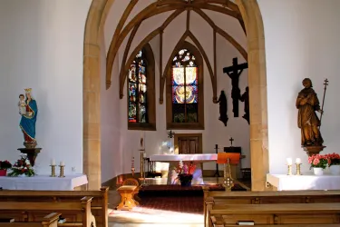 Eine kleine Kirche mit einer hohen Decke und einem Altar. Bunte Fenster und Statuen von Heiligen sind im Raum sichtbar.