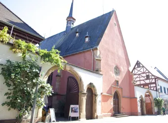 Ein historisches Gebäude mit roter Fassade und einem hohen Dach. Neben dem Gebäude befinden sich grüne Pflanzen und ein traditionelles Fachwerkhaus.