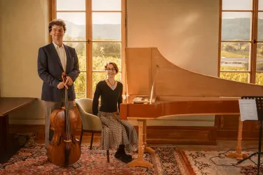Zwei Musiker stehen in einem hellen Raum mit großen Fenstern. Einer hat ein Cello, während die andere an einem Klavier sitzt.