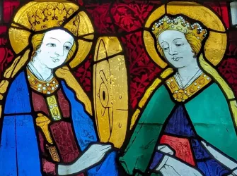 Ein buntes Glasfenster mit zwei Heiligen in historischen Gewändern. Die Figuren stehen einander gegenüber und haben goldene Heiligenscheine.