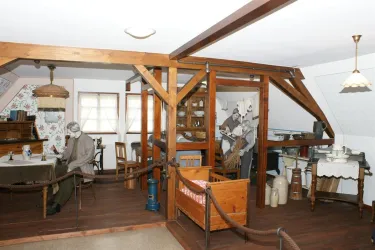 Ein traditionell eingerichteter Raum mit Holzdekor und einem Tisch. Eine Figur sitzt am Tisch, umgeben von alten Haushaltsgegenständen.