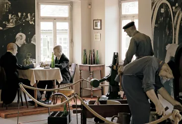 Ein historisches Café mit Wachsfiguren, die an einem Tisch sitzen und eine Unterhaltung führen. Im Hintergrund sind Flaschen und Maschinen aus der Vergangenheit zu sehen.