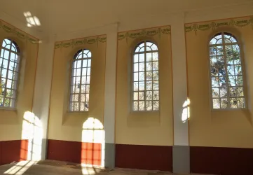 Ein heller Raum mit hohen Fenstern und sanften Lichtspielen. Die Wände sind in Pastellfarben gestrichen und zeigen eine einfache, aber ansprechende Dekoration.