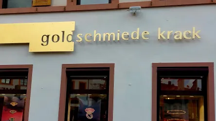 Das Bild zeigt ein Geschäftsschild mit dem Namen "goldschmiede krack". Im Hintergrund sind Fenster sichtbar, die zum Schaufenster des Geschäfts gehören.