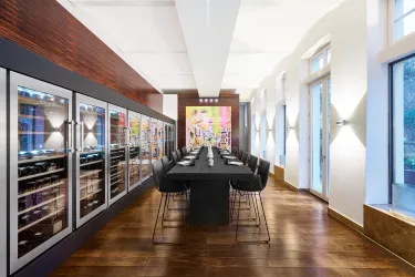 Ein eleganter Speisesaal mit einem langen Tisch und modernen Stühlen. An der Wand befinden sich große Weinschränke und im Hintergrund ein farbenfrohes Kunstwerk.