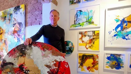 Ein Mann steht in einer Galerie und hält ein großes, buntes Kunstwerk. Um ihn herum hängen verschiedene Gemälde an der Wand.