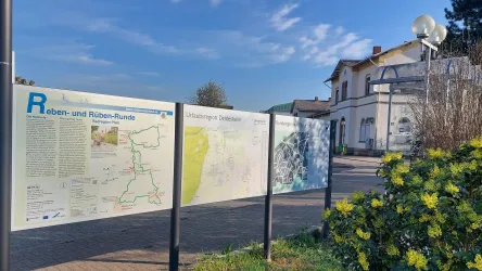 Eine Informationstafel mit Wander- und Radwegen ist in der Mitte des Bildes zu sehen. Im Hintergrund steht ein Gebäude und die Sonne scheint.