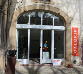Die Fassade einer Galerie mit großen Fenstern und einem Eingangsbereich. Ein Schild zeigt "offen" und es gibt eine bunte Skulptur im Inneren.