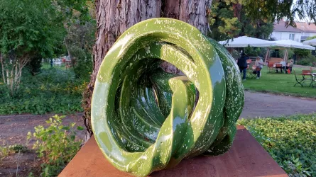 Eine grüne Skulptur mit spiralförmigen Formen steht auf einem Tisch im Freien. Im Hintergrund sind Bäume und eine Gartenlandschaft sichtbar.