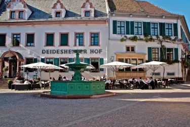 Ein malerischer Platz mit einem Brunnen in der Mitte und mehreren Tischen unter Sonnenschirmen. Im Hintergrund sind schöne Gebäude sichtbar, die eine einladende Atmosphäre schaffen.