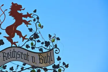 Ein kunstvoller Metallschild mit einem Löwen und Traubenmotiven. Darauf steht "Reichsrat d. Buhl" vor einem klaren blauen Himmel.