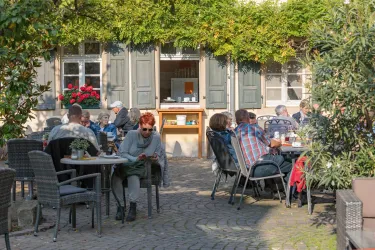 Ein gemütliches Café mit Außenplätzen, umgeben von Pflanzen. Gäste sitzen entspannt und genießen ihre Zeit im Freien.