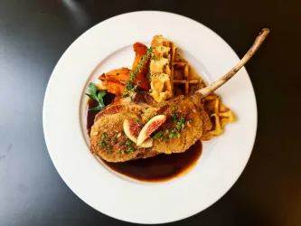 Ein Teller mit knusprigem Lammkotelett, serviert mit Waffeln und gebratenem Gemüse. Das Gericht ist mit frischen Kräutern und Feigen dekoriert.