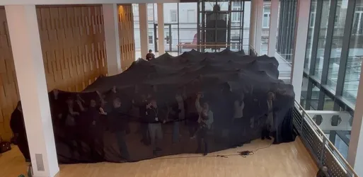 Ein großer schwarzer Vorhang in einem hellen Raum, der viele Menschen verdeckt. Die Stimmung wirkt geheimnisvoll und experimentell.