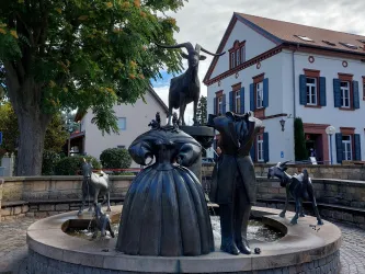 Eine kreative Brunnenskulptur mit Figuren von Menschen und Tieren, darunter eine Ziege und verschiedene andere Tiere. Der Brunnen steht vor einem historischen Gebäude in einer ruhigen Umgebung.