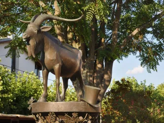 Eine bronzene Statue eines Ziegenbocks mit einem Zylinderhut. Sie steht vor einem grünen Hintergrund aus Bäumen und Blumen.