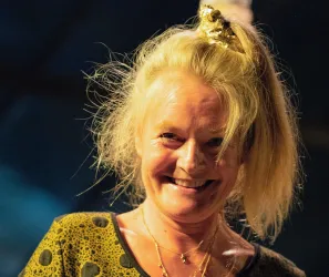 Eine lächelnde Frau mit zerzaustem Haar und einem goldenen Haaraccessoire. Sie trägt ein auffälliges, gepunktetes Oberteil.