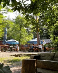 Eine einladende Gaststätte im Grünen mit Schatten von Bäumen. Gäste genießen ihre Zeit unter blauen Schirmen.