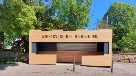 Ein modernes Gebäude mit dem Schriftzug "Winzerverein Deidesheim". Es befindet sich in einer grünen Umgebung mit Bäumen.