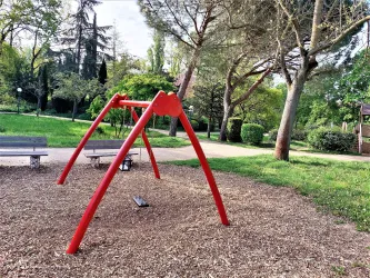 Ein Spielplatz mit einer roten Schaukel steht auf einem Holzhackschnitzelboden. Umgeben von Bäumen und einer grünen Wiese bietet der Ort eine einladende Atmosphäre.